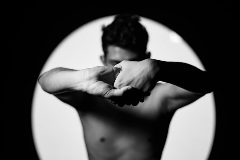 A black and white image of a shirtless man, blurred behind crossed arms in front. The pose evokes a shield or an internal struggle, capturing a sense of defense or emotional resistance.