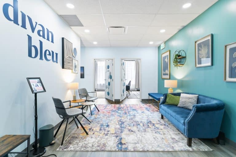 La salle d'attente de la clinique de thérapie au Divan bleu à Laval, affichant une atmosphère accueillante avec des murs teints de bleu jade, un canapé en velours bleu, des chaises modernes en tissu gris, une moquette aux teintes pastel, et une décoration murale artistique et contemporaine.