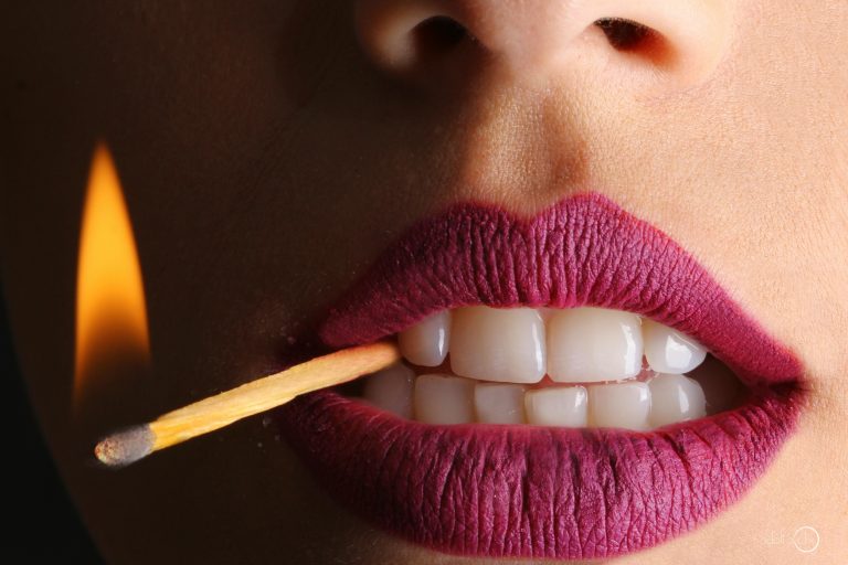 A close up of a womans mouth with bright pink lipstick on. She's biting down on a lit matchstick.