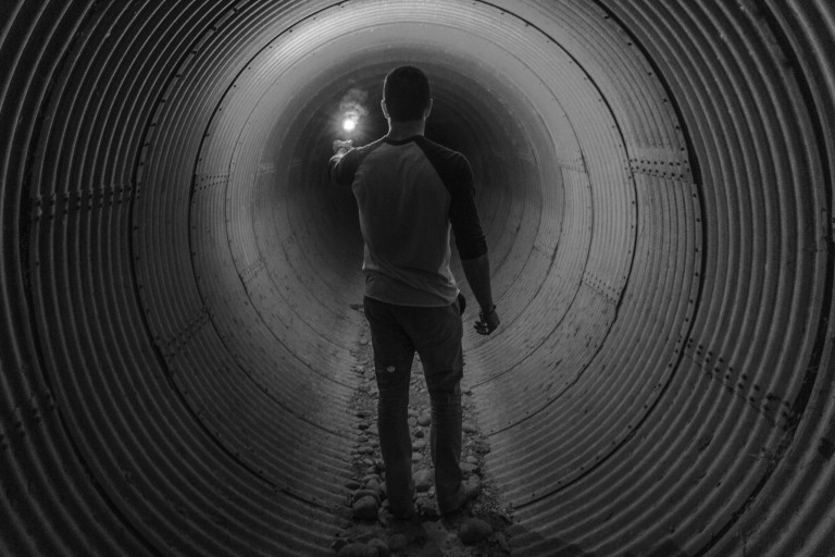 Silhouette d'un homme de dos éclairant l'obscurité d'un tunnel avec une torche. Cette image évoque le parcours individuel à travers les épreuves de la vie et la recherche de lumière face au trauma, un sujet profondément exploré dans le domaine de la santé mentale.
