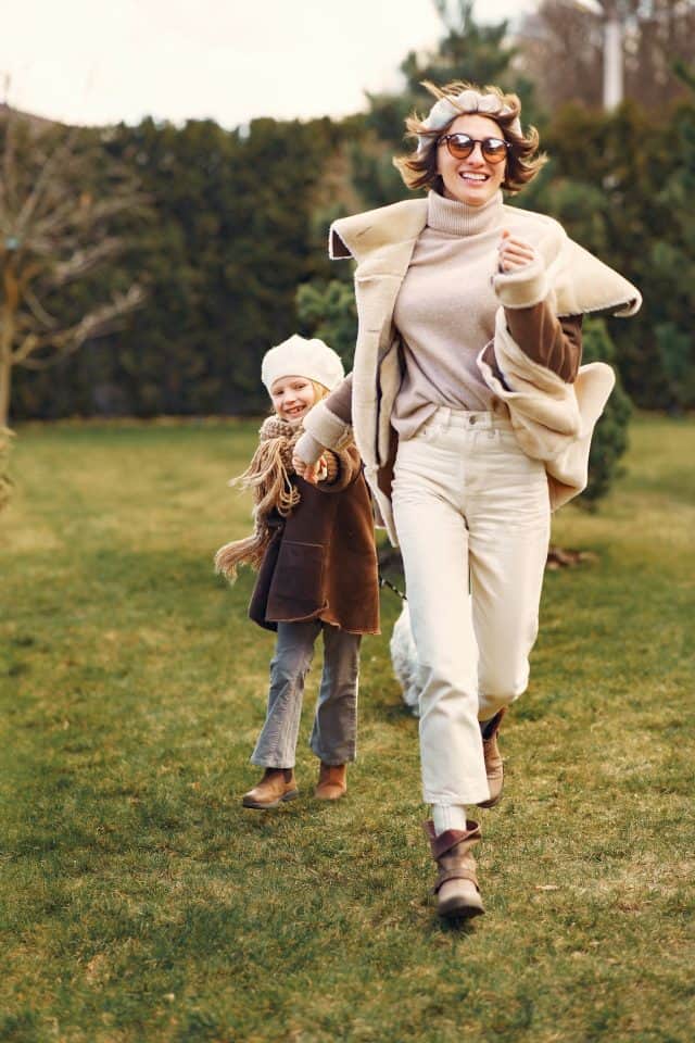 A smiling woman and a little girl with blond hair run on green grass under a cloudy blue sky. They look happy and carefree.